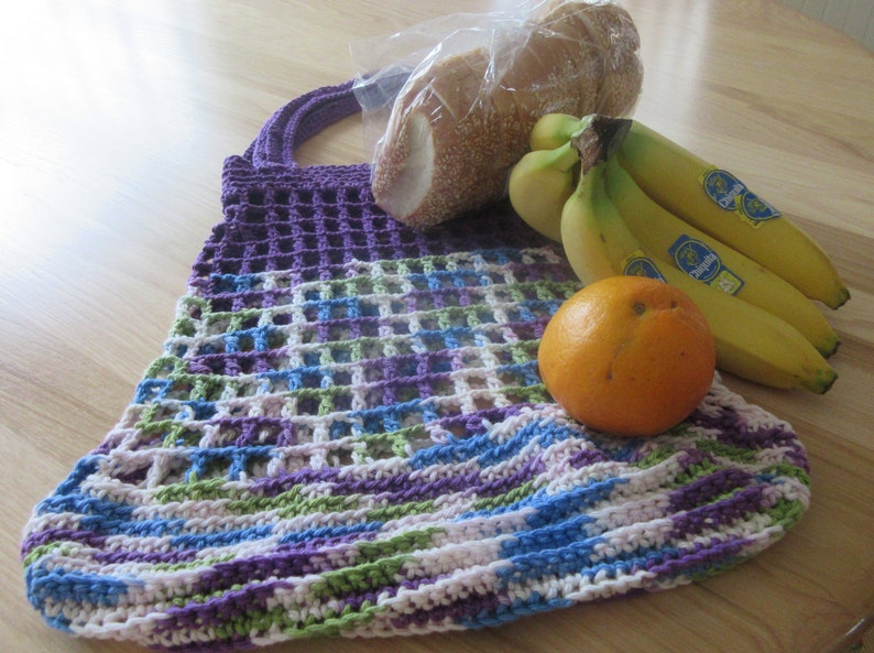May include: A purple, green, blue, and white crocheted mesh bag with a loaf of bread and bananas inside. An orange is sitting on top of the bag.