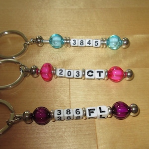 May include: Three metal keychains featuring silver-tone lobster claw clasps and split rings. Each keychain has a central segment of white square beads with black letters and numbers, flanked by two translucent colored beads and silver spacers.