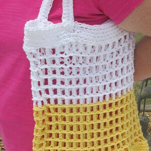 May include: A handmade crochet tote bag in white and yellow. The bag features a large open weave design with sturdy white handles. The bottom portion of the bag is yellow, transitioning to white at the top. The bag is held by a person.
