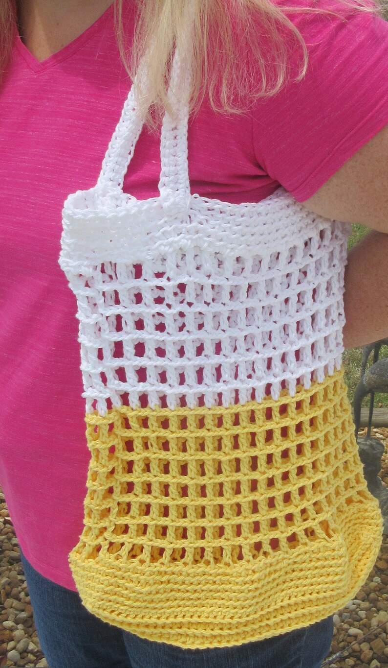 May include: A handmade crochet tote bag in white and yellow. The bag features a large open weave design with sturdy white handles. The bottom portion of the bag is yellow, transitioning to white at the top. The bag is held by a person.