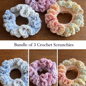 May include: A bundle of three crochet scrunchies in shades of blue, pink, and peach. The scrunchies are made of a soft, textured yarn and are arranged on a wooden surface. The text "Bundle of 3 Crochet Scrunchies" is also visible.