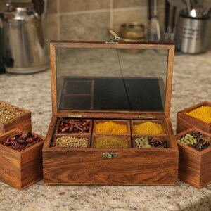 May include: A wooden spice box with a glass lid, open to reveal six compartments filled with various spices. Four small wooden boxes filled with spices are arranged around the main box. The spices are in shades of red, yellow, and green.