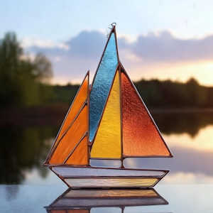 Könnte beinhalten: Ein Buntglas-Segelboot mit farbenfrohem Design. Die Segel sind orange, gelb, blau und rot, mit einem weißen Rumpf und einer reflektierenden Oberfläche. Der Hintergrund zeigt einen See und einen Sonnenuntergang.
