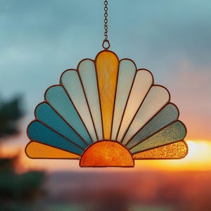 May include: A stained glass suncatcher in a fan shape, with a half-circle base and radiating segments in shades of blue, yellow, and white. The suncatcher is suspended by a chain, with a sunset in the background.