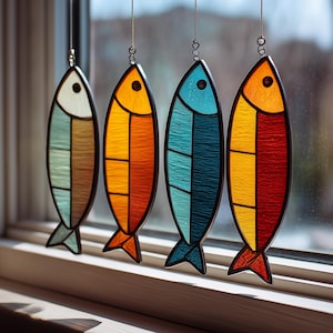 May include: Four colorful stained glass fish hang in front of a window. Each fish has a black outline and is divided into sections of color, including blue, orange, yellow, and red. The fish are suspended by thin wires.