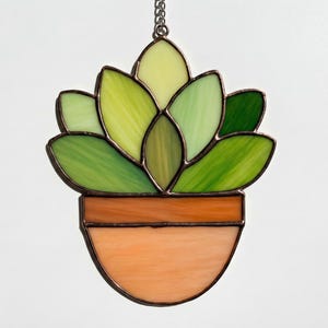 Puede incluir: Un atrapasoles de vidrieras con forma de planta suculenta en una maceta color terracota. La planta presenta hojas de vidrio en varios tonos de verde con soldaduras de metal oscuro, y la maceta es de vidrio naranja claro con una línea.