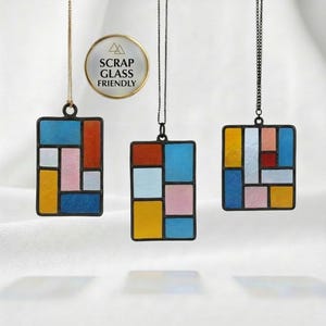 May include: Three stained glass pendant necklaces with geometric designs in shades of blue, orange, pink, and yellow. The pendants are rectangular and hang from delicate chains. A circular emblem reads "SCRAP GLASS FRIENDLY".
