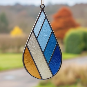 May include: A stained glass teardrop-shaped suncatcher with a black metal frame. The design features sections of orange, cream, and various shades of blue. A small loop at the top allows for hanging. The background is blurred, showing a natural outdoor scene.