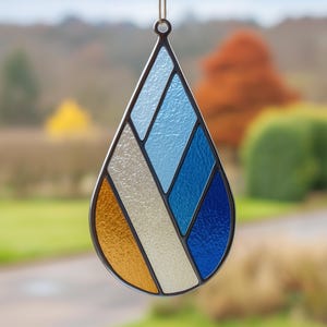 May include: A stained glass teardrop-shaped suncatcher with a black metal frame. The design features sections of orange, cream, and various shades of blue. A small loop at the top allows for hanging. The background is blurred, showing a natural outdoor scene.
