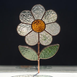 May include: A stained glass daisy with a copper stem and leaves. The flower has a yellow center and clear petals with textured patterns. The leaves are green, and the stem is copper-colored. The flower is set against a dark background.