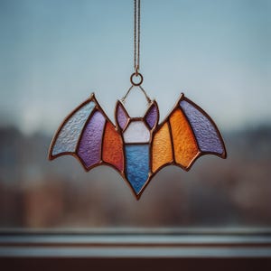 Puede incluir: Un murciélago de vidriera con un marco de cobre y una cadena para colgar. Las alas del murciélago están divididas en secciones de vidrio azul, morado y naranja. El cuerpo es blanco, azul y naranja.