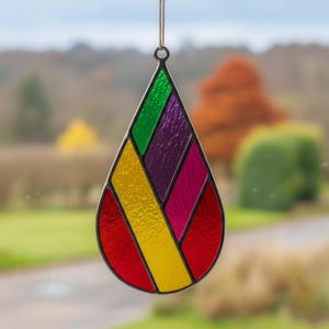 Pode incluir: Enfeite em forma de gota de vidro manchado com um design geom&eacute;trico. Apresenta sec&ccedil;&otilde;es de vidro vermelho, amarelo, verde, roxo e rosa, delineadas em preto. Inclui uma pequena argola de metal e cord&atilde;o para pendurar.