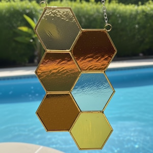 May include: A stained glass honeycomb suncatcher with six hexagonal panels in shades of gold, amber, and blue. The panels are framed in gold metal and suspended by a chain. The background shows a pool and greenery.