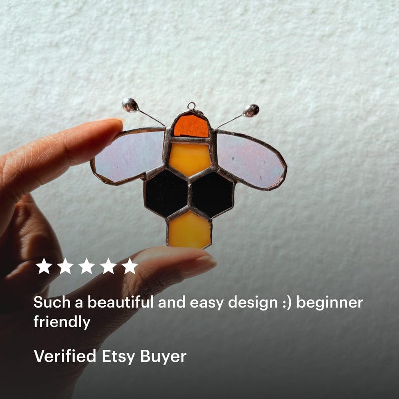 May include: A stained glass bee ornament with a honeycomb design. The bee features orange, yellow, black, and white glass pieces, with wire antennae. The text reads: "Such a beautiful and easy design :) beginner friendly".