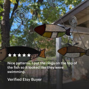 May include: Three stained glass fish ornaments in shades of red, yellow, and brown hang from chains attached to suction cups. The fish have segmented bodies and are suspended in front of a window with a blurred outdoor background.