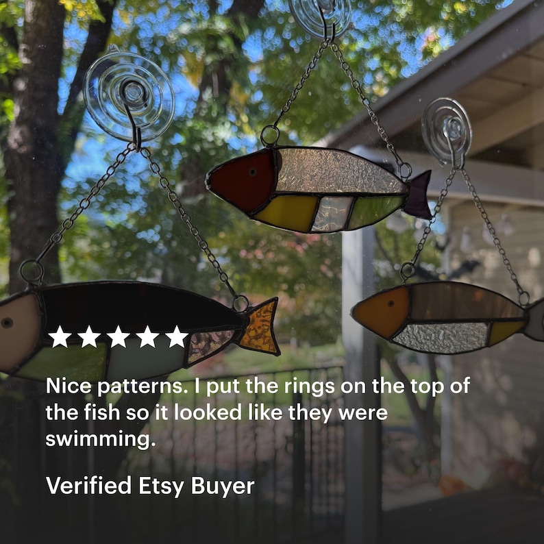 May include: Three stained glass fish ornaments in shades of red, yellow, and brown hang from chains attached to suction cups. The fish have segmented bodies and are suspended in front of a window with a blurred outdoor background.