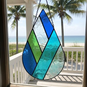 Pode incluir: Uma janela suspensa em forma de l&aacute;grima de vitral, com se&ccedil;&otilde;es de vidro azul e verde. A janela &eacute; suspensa por uma corrente e pendurada em frente a uma cena de praia com palmeiras.
