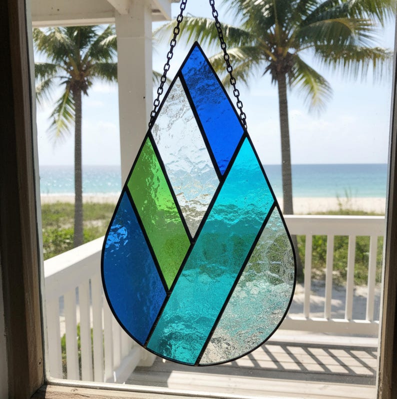 Pode incluir: Uma janela suspensa em forma de l&aacute;grima de vitral, com se&ccedil;&otilde;es de vidro azul e verde. A janela &eacute; suspensa por uma corrente e pendurada em frente a uma cena de praia com palmeiras.