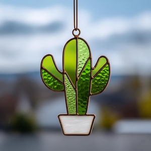 May include: A stained glass cactus suncatcher with a copper-colored frame. The cactus is various shades of green, and the pot is white. The suncatcher hangs from a thin, brown cord. The background is blurred, with a view of a cloudy sky.