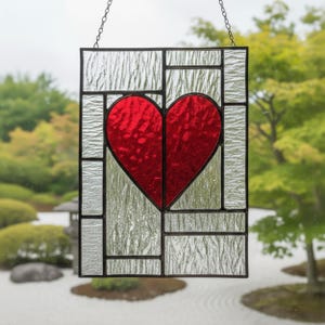 Op de afbeelding: Gebrandschilderd glaspaneel met een rood hart in het midden, omgeven door helder, getextureerd glas. Het zwart omlijste paneel hangt aan een ketting. Geschikt voor raamdecoratie, ongeveer 20 cm x 30 cm.