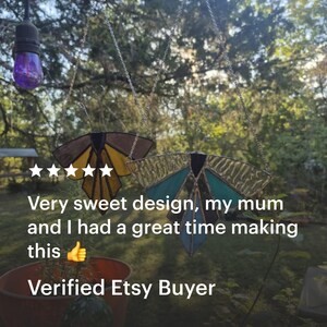 Pode incluir: Dois ornamentos de borboleta em vitral pendurados em correntes. Uma borboleta tem asas castanhas e amarelas, a outra tem asas turquesas e douradas. O texto diz: "Very sweet design, my mum and I had a great time making this. Verified Etsy Buyer."
