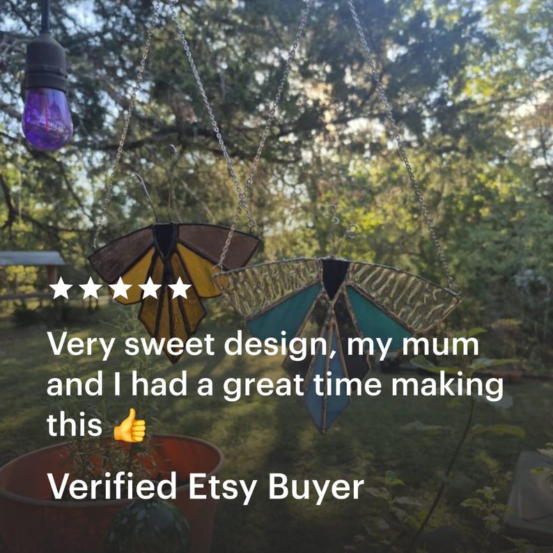 Pode incluir: Dois ornamentos de borboleta em vitral pendurados em correntes. Uma borboleta tem asas castanhas e amarelas, a outra tem asas turquesas e douradas. O texto diz: "Very sweet design, my mum and I had a great time making this. Verified Etsy Buyer."