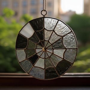 May include: A circular stained glass suncatcher featuring a spiderweb pattern. The glass segments are in white, grey, and black, set within a gold-toned metal frame. It has a small chain for hanging. The suncatcher is shown against a blurred green backdrop.
