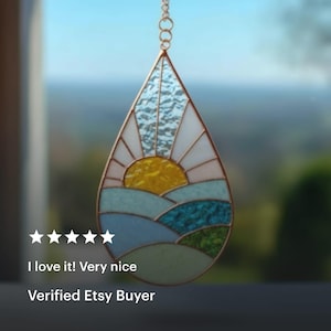 May include: Stained glass suncatcher in a teardrop shape. The design features a yellow sun, white rays, and blue and green landscape elements. The suncatcher is suspended by a gold chain. The text "I love it! Very nice" and "Verified Etsy Buyer" are visible.