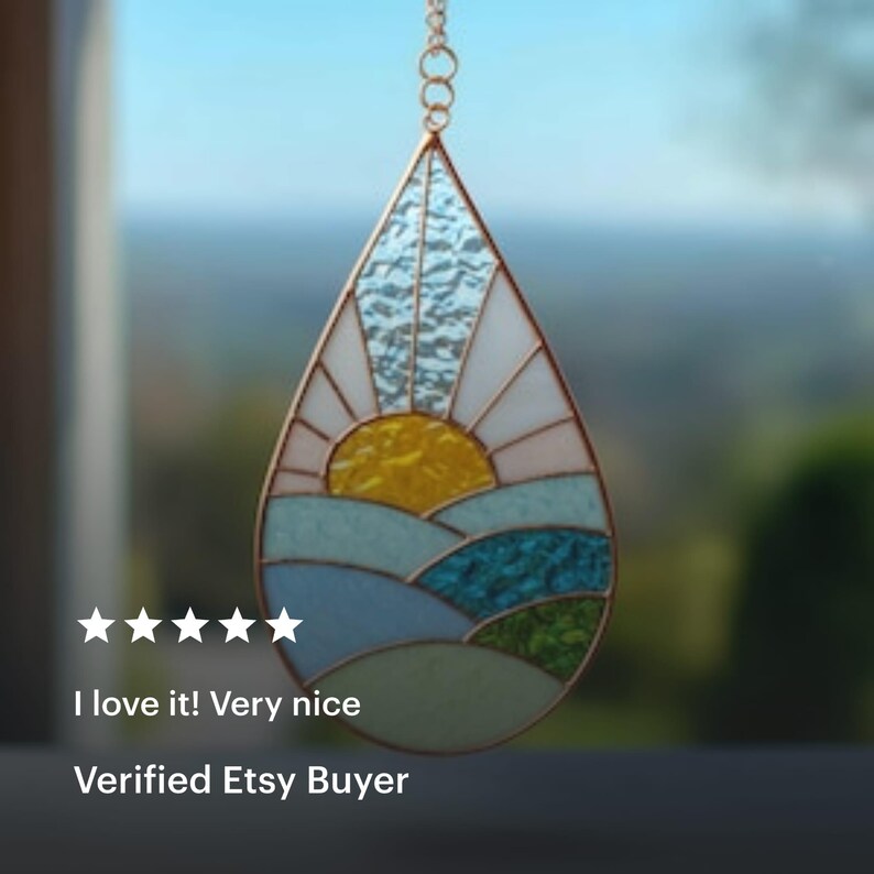 May include: Stained glass suncatcher in a teardrop shape. The design features a yellow sun, white rays, and blue and green landscape elements. The suncatcher is suspended by a gold chain. The text "I love it! Very nice" and "Verified Etsy Buyer" are visible.