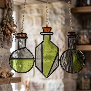 Può includere: Tre bottiglie di pozione in vetro colorato appese a catene dorate. Le bottiglie, di varie forme, contengono liquidi verdi e trasparenti e sono sigillate con tappi di sughero marrone. Il design include forme rotonde e geometriche.