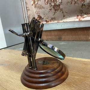 May include: A vintage-style magnifying glass with a dark metal frame and a wooden base. The magnifying glass is mounted on an adjustable arm. The base is round and has a tiered design. The background features a floral pattern.