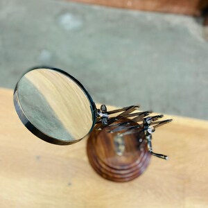May include: A magnifying glass with a round lens and a dark metal frame. The lens is positioned above a wooden base with a decorative metal dragonfly design. The base is made of dark wood.