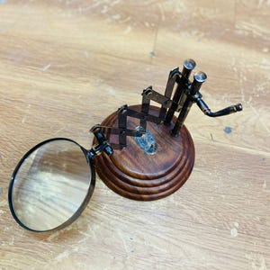 May include: A magnifying glass with a dark metal frame and a round lens is attached to a wooden base. The base has a tiered design and a dark brown finish. The magnifying glass is positioned to the left, with the lens facing the viewer.
