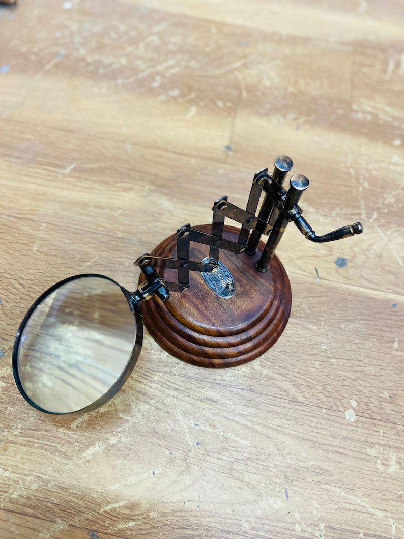 May include: A magnifying glass with a dark metal frame and a round lens is attached to a wooden base. The base has a tiered design and a dark brown finish. The magnifying glass is positioned to the left, with the lens facing the viewer.