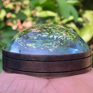 May include: A circular paperweight featuring a smooth, clear glass dome top that reflects green foliage. The base is constructed from a dark, brushed metallic material with two horizontal grooves encircling the cylindrical side profile.
