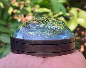 May include: A circular paperweight featuring a smooth, clear glass dome top that reflects green foliage. The base is constructed from a dark, brushed metallic material with two horizontal grooves encircling the cylindrical side profile.