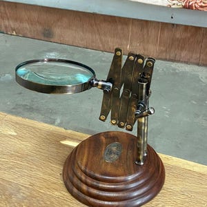 May include: A vintage-style magnifying glass with a round lens and an adjustable brass arm. The lens is set in a dark metal frame. The base is made of dark wood with a circular design. A small metal emblem is affixed to the wooden base.