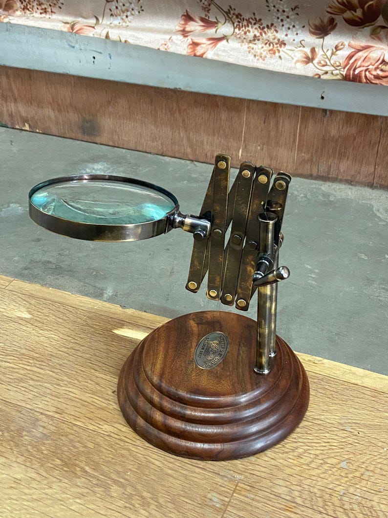 May include: A vintage-style magnifying glass with a round lens and an adjustable brass arm. The lens is set in a dark metal frame. The base is made of dark wood with a circular design. A small metal emblem is affixed to the wooden base.