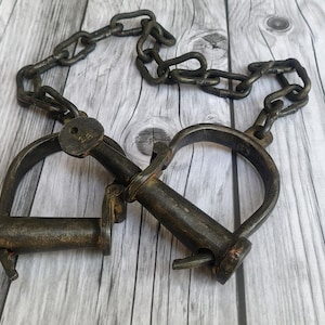 Peut inclure: Une paire de menottes en métal sombre et patiné, reliées par une courte chaîne robuste. Les menottes présentent une forme arrondie avec un mécanisme de verrouillage et une barre cylindrique, montrant des signes d'oxydation et d'usure.
