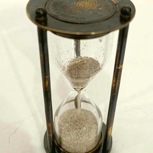 May include: An antique hourglass with a dark metal frame and clear glass bulbs. The top of the hourglass has the words "Ross London Sand Timer" engraved. The sand is a light tan color.