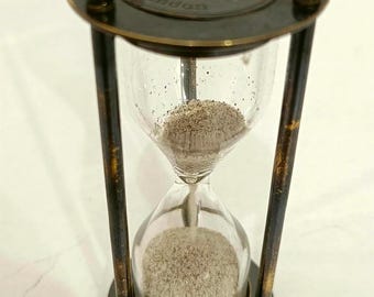 May include: An antique hourglass with a dark metal frame and clear glass bulbs. The top of the hourglass has the words "Ross London Sand Timer" engraved. The sand is a light tan color.