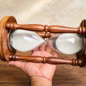 Puede incluir: Un reloj de arena horizontal con un marco de madera marrón pulida, tapas circulares en los extremos y cuatro pilares de soporte torneados. Los bulbos de vidrio transparente contienen arena blanca fina que fluye entre las cámaras.