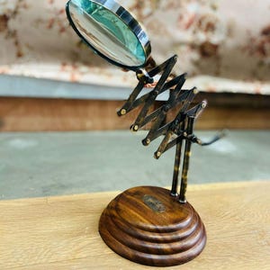 May include: A vintage-style magnifying glass with a brass frame and a wooden base. The magnifying glass has an adjustable scissor arm and a round lens. The base is made of dark wood with a decorative design. The magnifying glass is a decorative desk accessory.