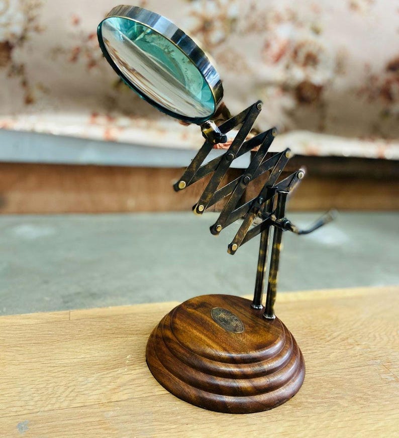 May include: A vintage-style magnifying glass with a brass frame and a wooden base. The magnifying glass has an adjustable scissor arm and a round lens. The base is made of dark wood with a decorative design. The magnifying glass is a decorative desk accessory.