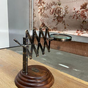 May include: A vintage-style magnifying glass with a brass-colored adjustable arm and a round glass lens. The base is made of dark wood with a circular design. The magnifying glass is extended, showing its full length. The background includes a floral patterned curtain.