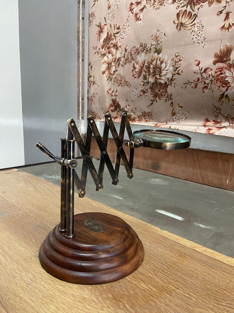 May include: A vintage-style magnifying glass with a brass-colored adjustable arm and a round glass lens. The base is made of dark wood with a circular design. The magnifying glass is extended, showing its full length. The background includes a floral patterned curtain.