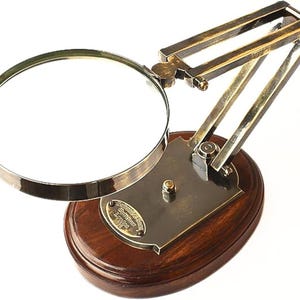 May include: A vintage-style magnifying glass with a brass-colored frame and adjustable arm, mounted on a dark wooden base. The magnifying glass has a circular lens and a small brass plate with text. The base is oval-shaped.