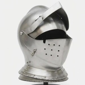 May include: A polished silver-colored metal knight helmet featuring a rounded dome, a hinged visor with a horizontal eye slit, and a lower face guard with a grid of small circular ventilation holes, all mounted on a circular black base.