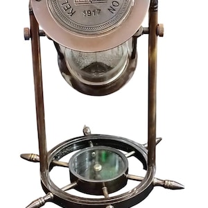 Puede incluir: Instrumento náutico antiguo de latón con brújula y reloj de arena. La parte superior tiene el texto "KELVIN & HUGHES LONDON 1917" alrededor de un emblema de corona. La base tiene un diseño de rueda de barco.