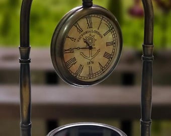 May include: A brass-toned metal desk clock and compass set featuring an arched stand. The clock face displays Roman numerals, the text "VICTORIA LONDON" and "1876", and a central anchor graphic. A circular magnetic compass is integrated into the base.